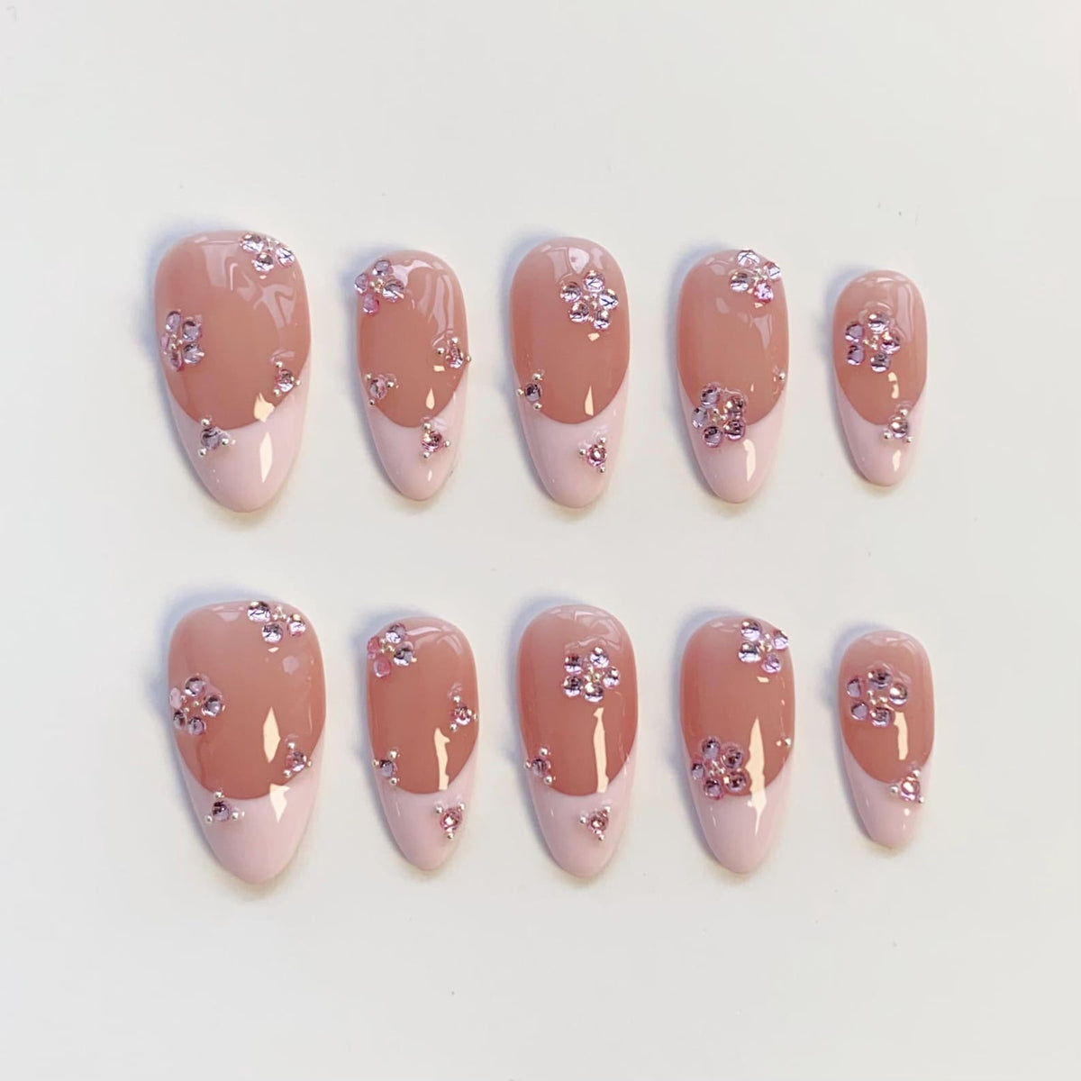 Set of ten artificial nails. Each nail features a light pink base with a darker pink tip. The nails are adorned with small, sparkling, pink floral embellishments. The nails are oval-shaped.
