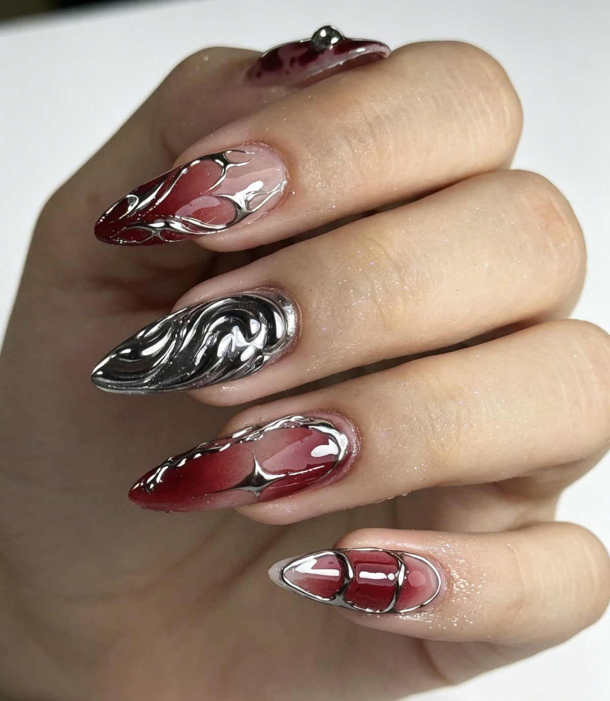 Almond-shaped artificial nails featuring a gradient of deep red and clear tones. The designs include raised, metallic silver liquid-style accents, swirling patterns, and a four-pointed star shape layered over the red base color.