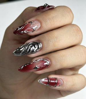 Almond-shaped artificial nails featuring a gradient of deep red and clear tones. The designs include raised, metallic silver liquid-style accents, swirling patterns, and a four-pointed star shape layered over the red base color.