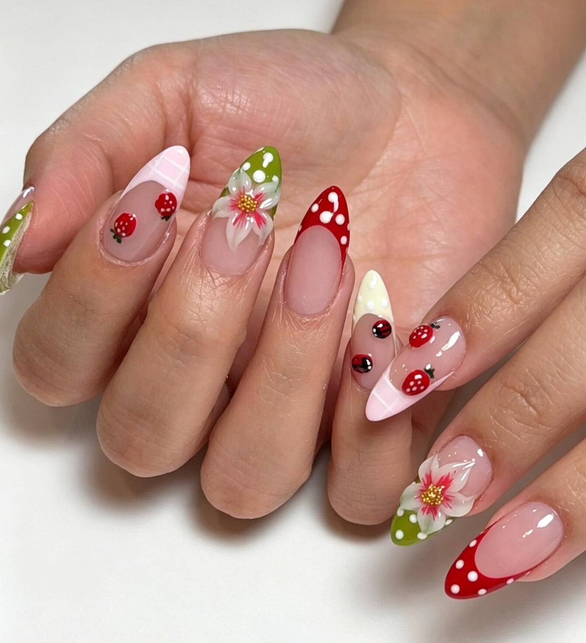 Almond-shaped nails featuring a French manicure style with varied designs. Some nails have red strawberry motifs, others display white floral patterns on green backgrounds, and some feature red or pink tips with white polka dots.