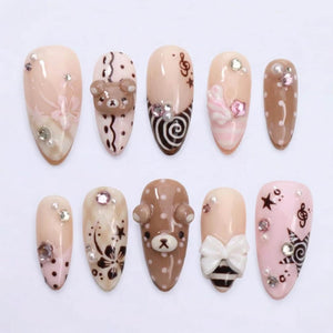 A set of ten almond-shaped press-on nails in shades of beige, brown, and pink. The designs feature 3D bear faces, white bows, floral patterns, musical notes, stars, polka dots, and swirls, accented with small clear and pink rhinestones.