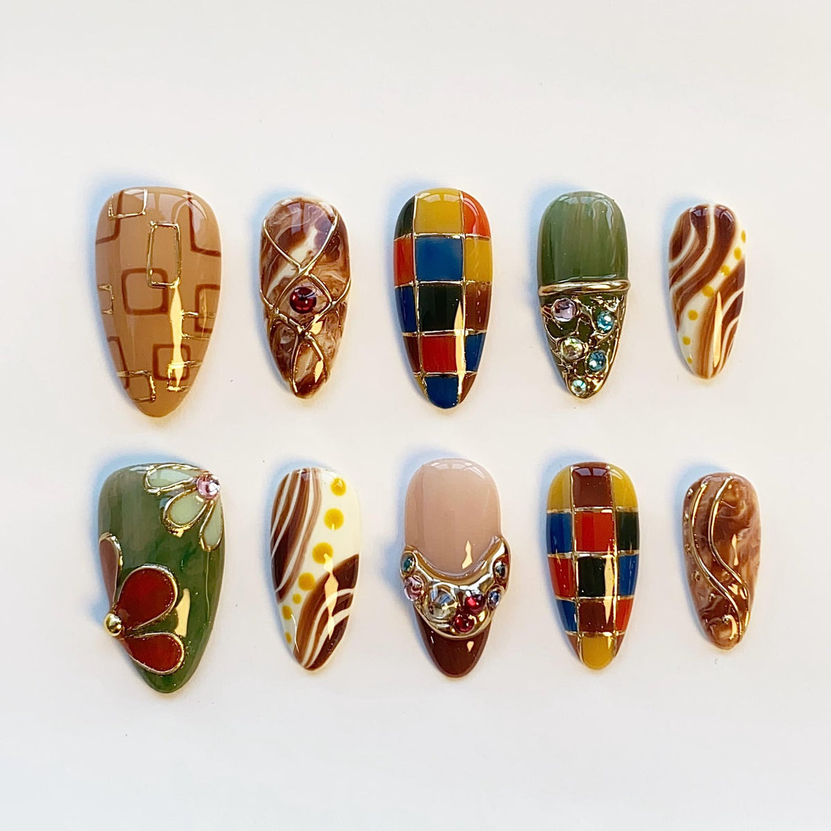 A set of ten almond-shaped press-on nails featuring diverse designs including gold geometric lines, marble swirls, multicolored checkered patterns, floral motifs, and embedded rhinestones in shades of brown, green, blue, and red.