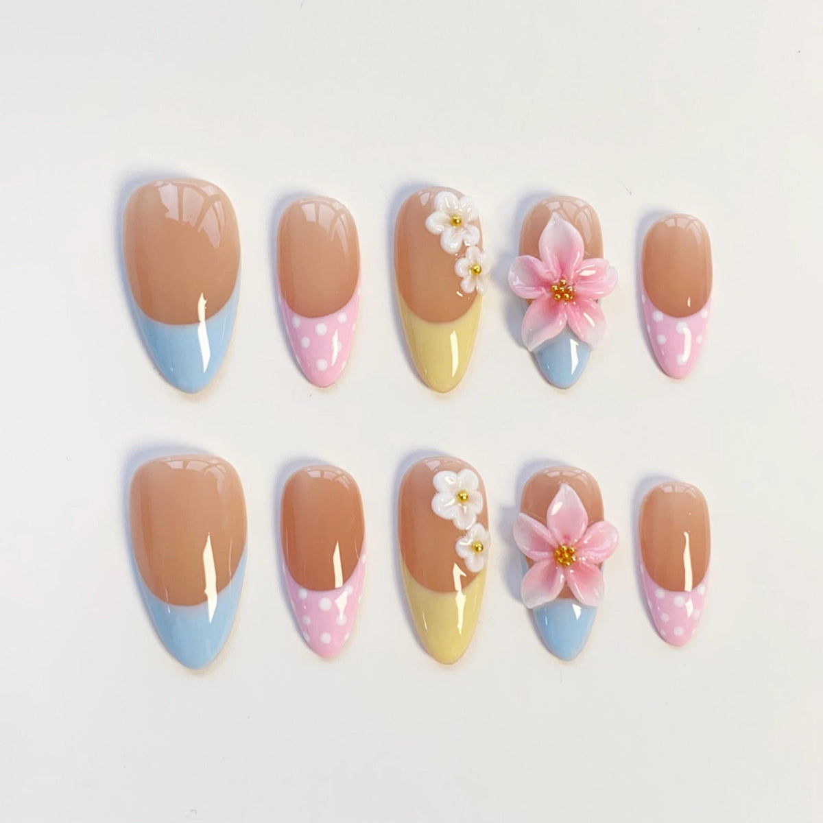 A set of ten almond-shaped press-on nails featuring a nude base with various tip designs. Some tips are solid light blue or yellow, while others are pink with white polka dots. Two nails feature 3D white flowers and pink flowers.