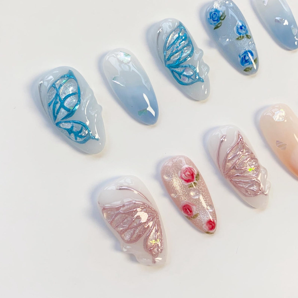 A set of almond-shaped press-on nails featuring blue and pink color palettes. Designs include raised 3D butterfly wings in metallic blue or pink, hand-painted blue roses, pink rosebuds, and iridescent flakes on a glossy finish.