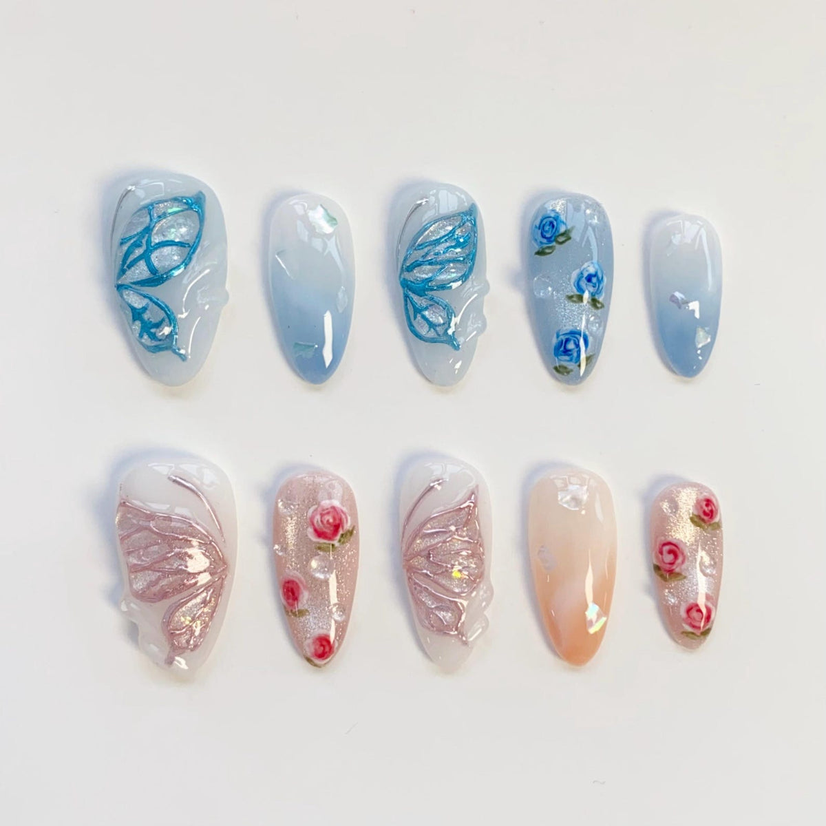 A set of ten press-on nails featuring almond-shaped tips. The top row displays blue butterfly wing designs and blue rose patterns on a white and blue gradient base. The bottom row features pink butterfly wings and pink rose motifs.