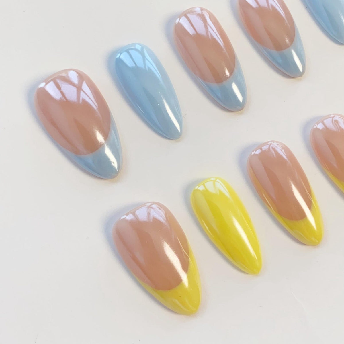 A set of almond-shaped press-on nails featuring a glossy finish. Some nails have a nude base with light blue tips, while others have a nude base with bright yellow tips. The nails are arranged in a staggered pattern on a white surface.