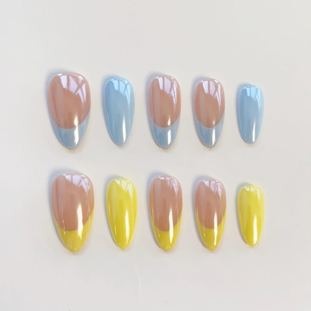 A set of ten almond-shaped press-on nails arranged in two rows. The top row features a nude base with light blue tips, while the bottom row features a nude base with bright yellow tips. Each nail has a smooth, glossy finish.