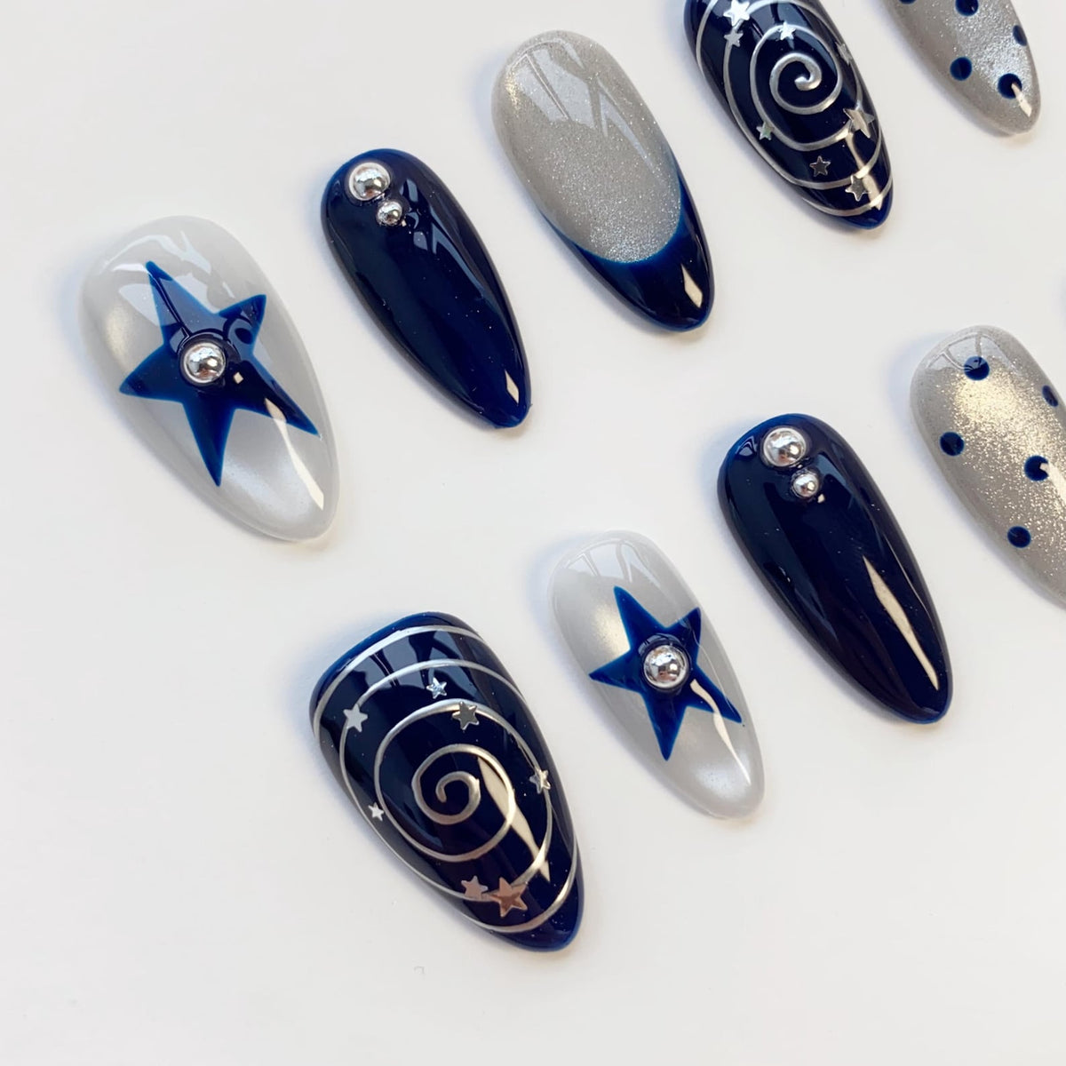A set of almond-shaped press-on nails featuring a celestial theme with deep navy blue and shimmering silver polish. Designs include blue stars with silver studs, silver spiral galaxies with tiny stars, and silver dots on a grey base.