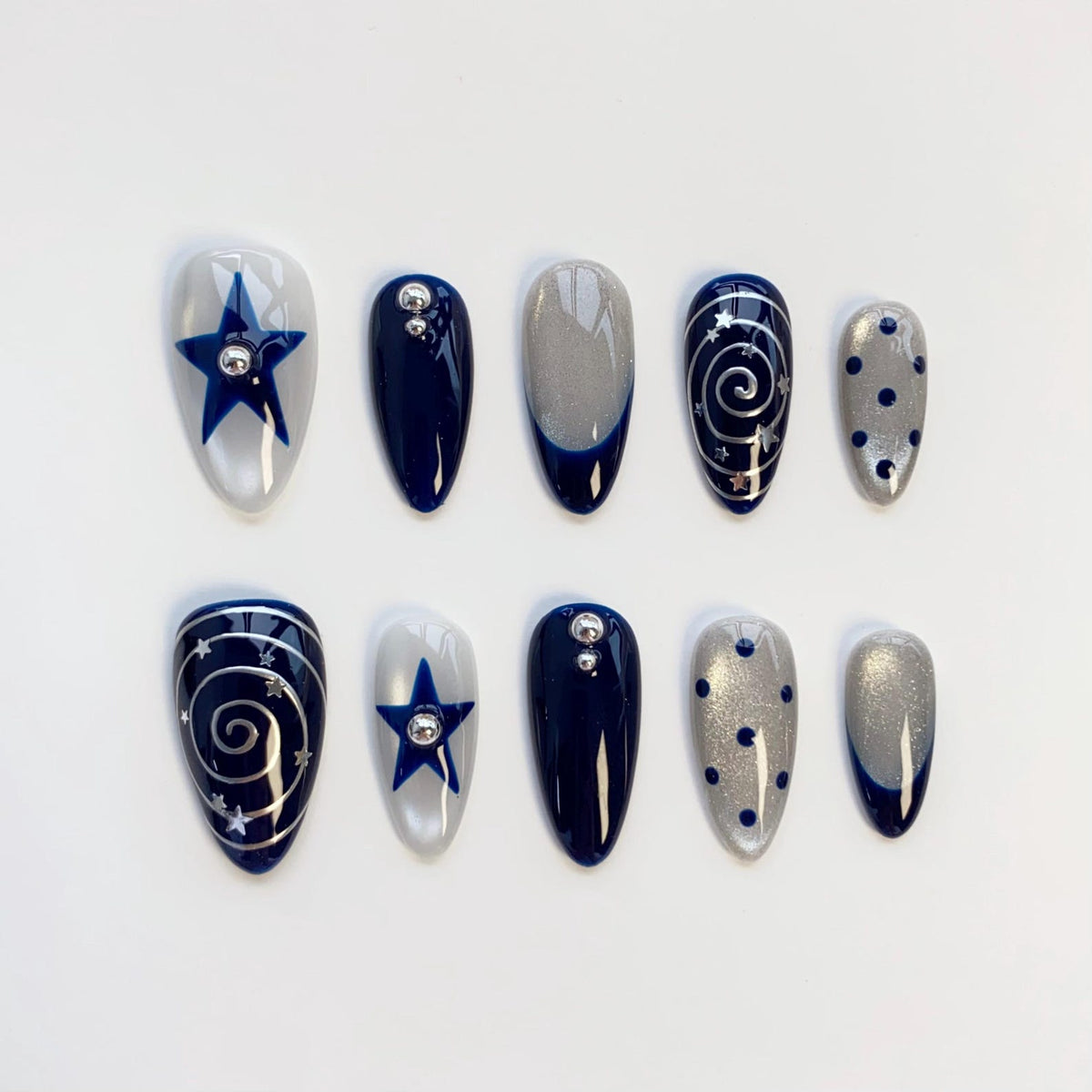 A set of ten almond-shaped press-on nails featuring a navy blue and silver color palette. Designs include solid navy blue, silver glitter with navy tips, navy blue stars, silver swirls with tiny stars, and navy blue polka dots.