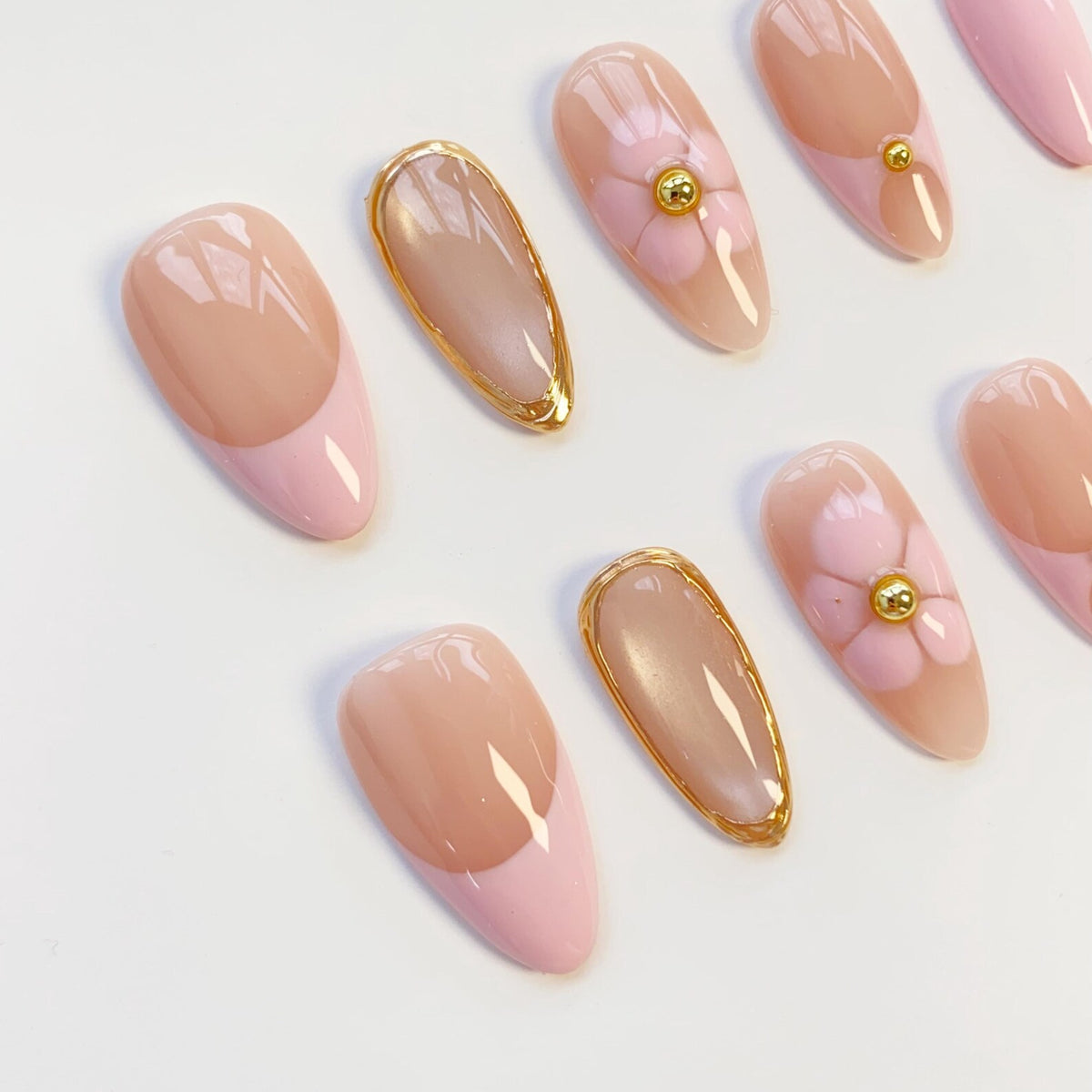 A set of almond-shaped press-on nails featuring a nude base with light pink tips. Some nails display a hand-painted light pink flower with a small gold metallic bead at the center, while others are outlined with a thin gold metallic border.