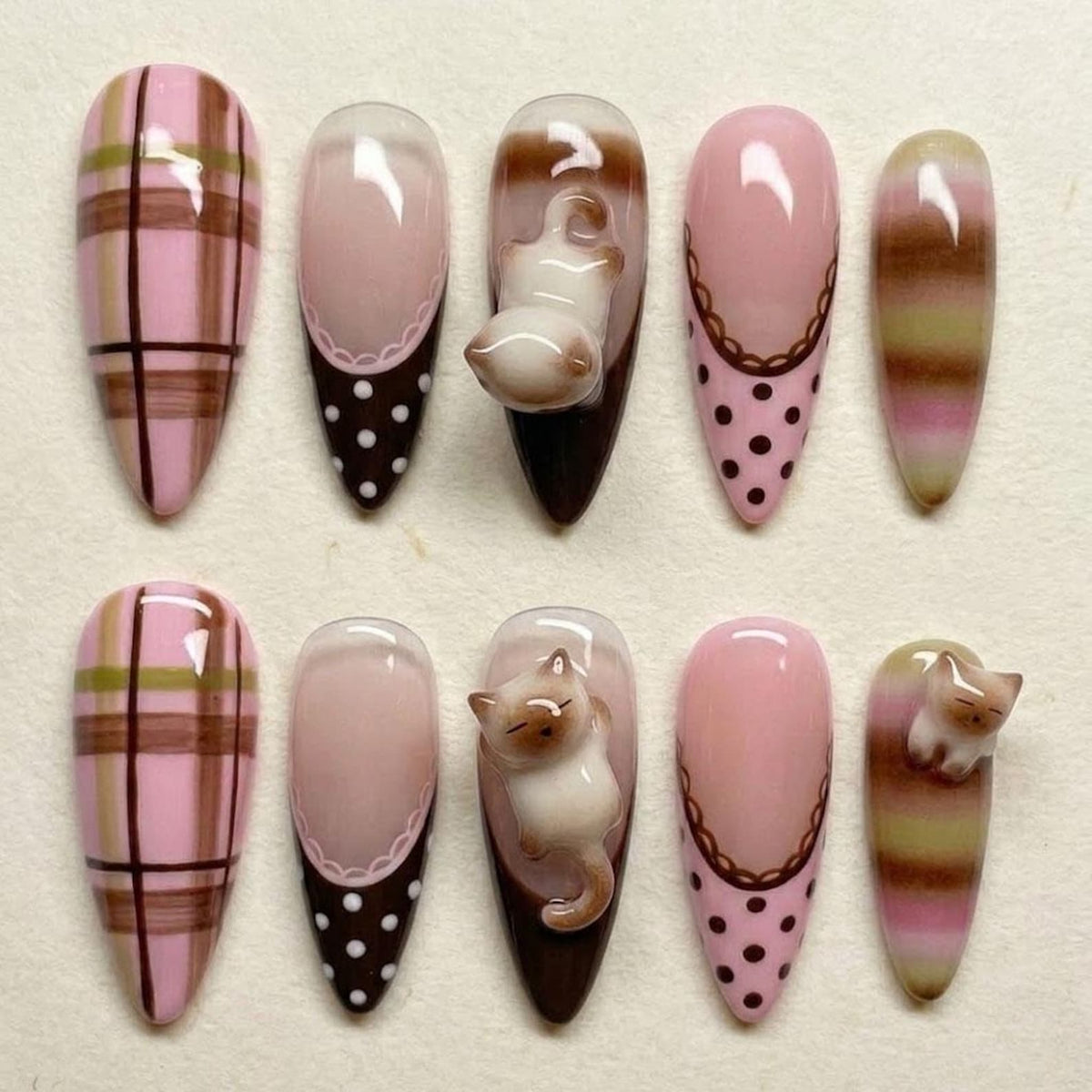 A set of ten almond-shaped false nails. Designs include pink plaid, solid pink with a white lace edge and brown polka dots, a 3D cat, and pink with a brown lace edge and brown polka dots. The nails are painted in shades of pink, brown, and white.