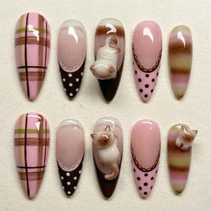 A set of ten almond-shaped false nails. Designs include pink plaid, solid pink with a white lace edge and brown polka dots, a 3D cat, and pink with a brown lace edge and brown polka dots. The nails are painted in shades of pink, brown, and white.