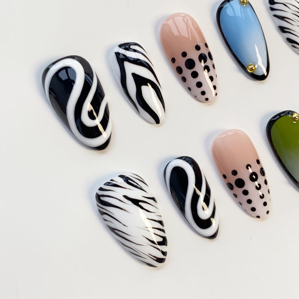 A collection of artificial nails with various designs. Some nails feature black and white swirls, others have zebra stripes, and some are painted with a nude base and black polka dots. A few nails have a blue or green gradient.