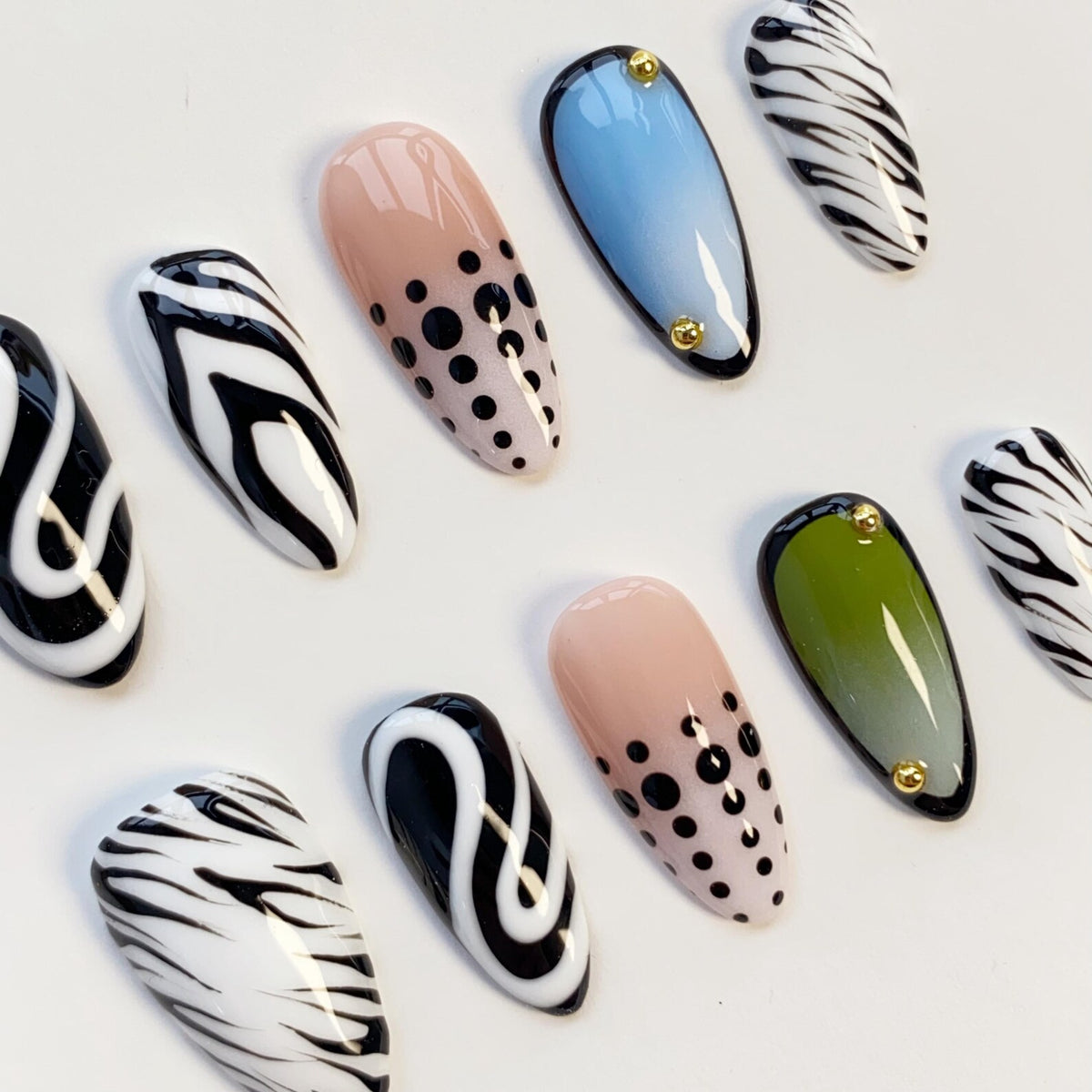 A collection of artificial nails in various designs. Some nails feature a black and white zebra stripe pattern, while others have a nude base with black polka dots. Two nails are light blue and olive green with gold accents.