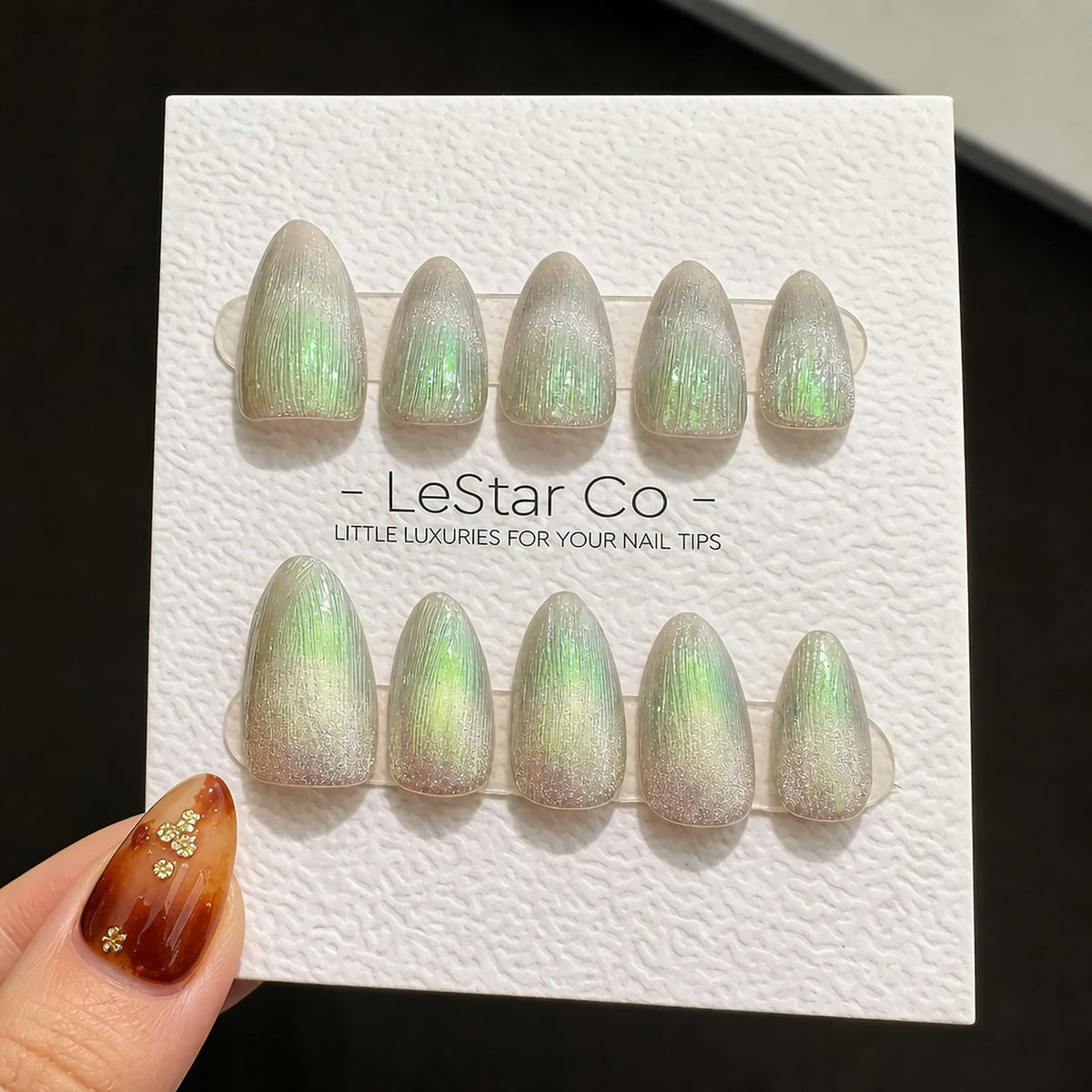 A set of ten almond-shaped press-on nails with a shimmering green and silver cat-eye effect, displayed on a white card labeled LeStar Co. LITTLE LUXURIES FOR YOUR NAIL TIPS. The nails feature a vertic