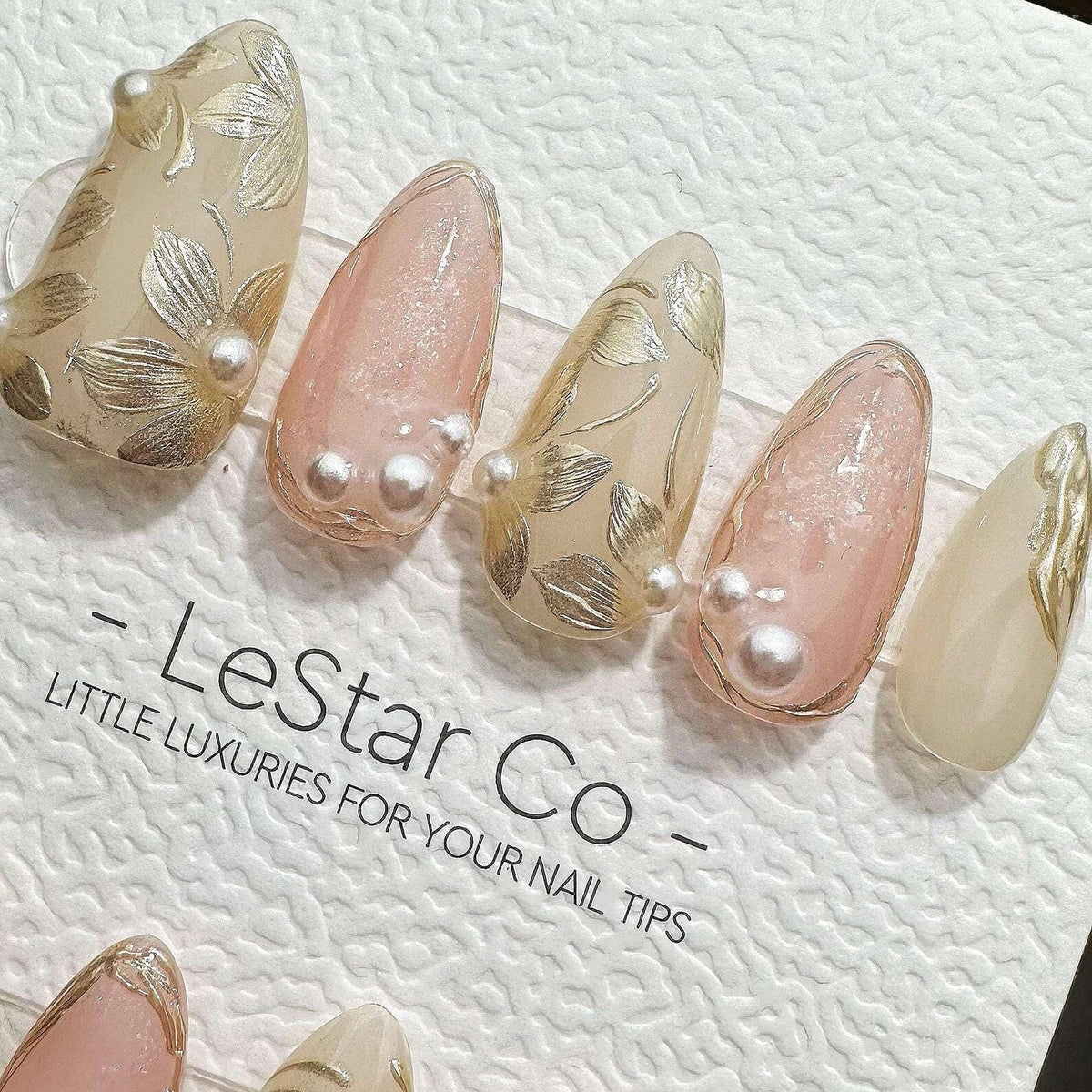 A set of almond-shaped press-on nails featuring a mix of nude and soft pink tones, decorated with metallic gold leaf floral patterns and small white pearl accents. The text LeStar Co - LITTLE LUXURIES