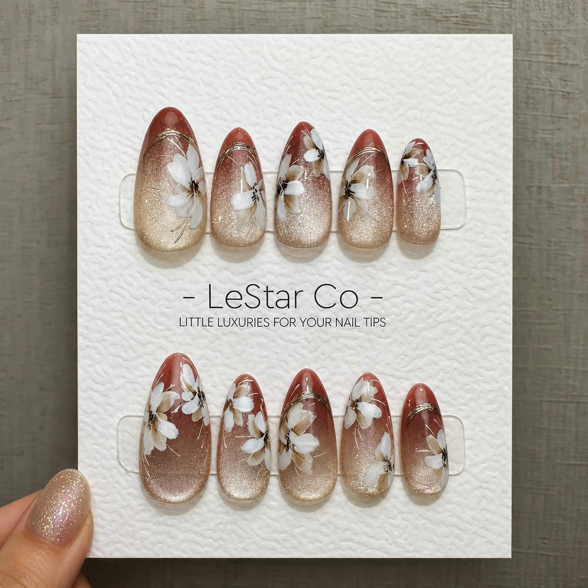 Set of almond-shaped press-on nails. Each nail features a gradient design, transitioning from a glittery gold base to a reddish-brown tip. Delicate white floral patterns adorn each nail, with the text