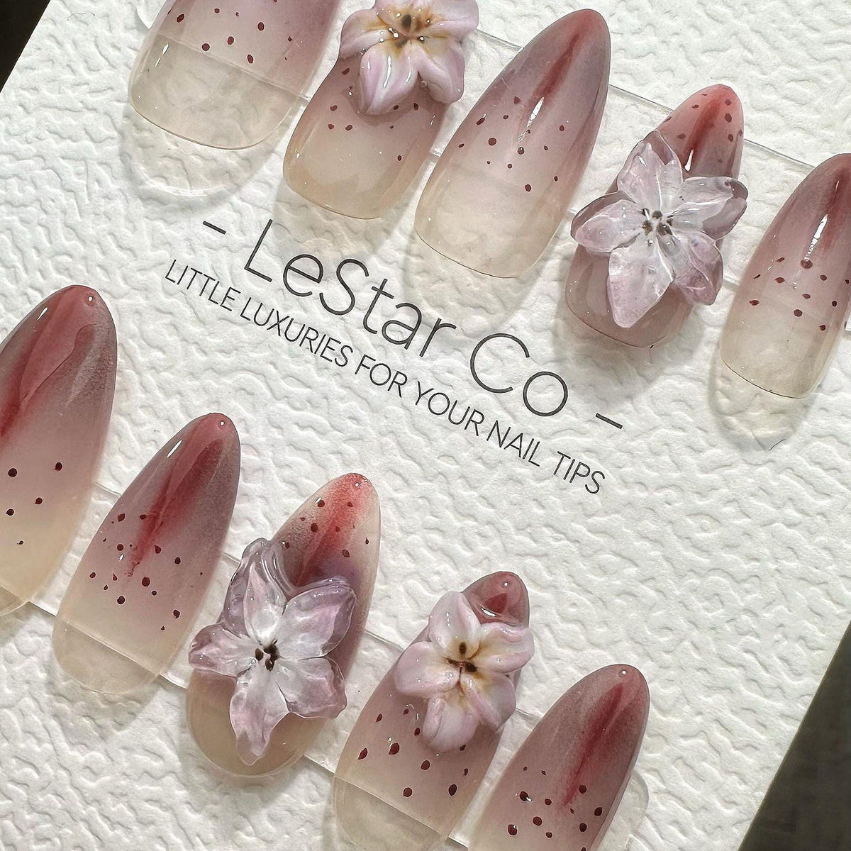 Oval-shaped nail tips with a gradient design, transitioning from a nude base to a reddish-pink hue. Some nails feature small brown dots and delicate, translucent flower embellishments. The text "- Les