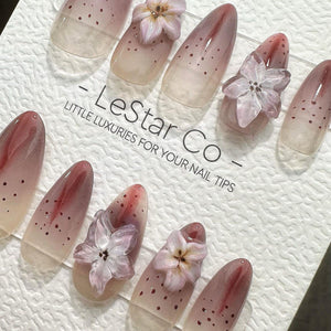 Oval-shaped nail tips with a gradient design, transitioning from a nude base to a reddish-pink hue. Some nails feature small brown dots and delicate, translucent flower embellishments. The text "- Les