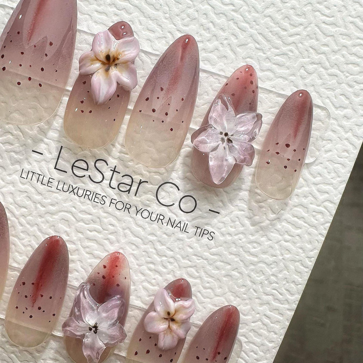 Oval-shaped artificial nails with a gradient design, transitioning from a nude base to a reddish-pink hue. Each nail features small dark dots and a delicate, three-dimensional flower. The text "- Lest
