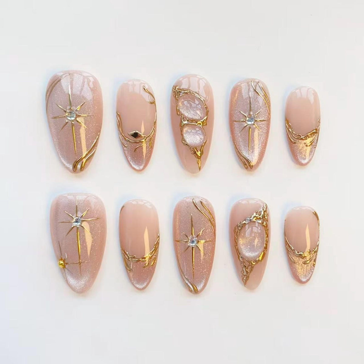 A set of ten almond-shaped false nails. Each nail is painted a pale pink with gold embellishments. Designs include starbursts, swirls, and ornate gold frames with clear, iridescent accents. The nails are arranged on a white surface.