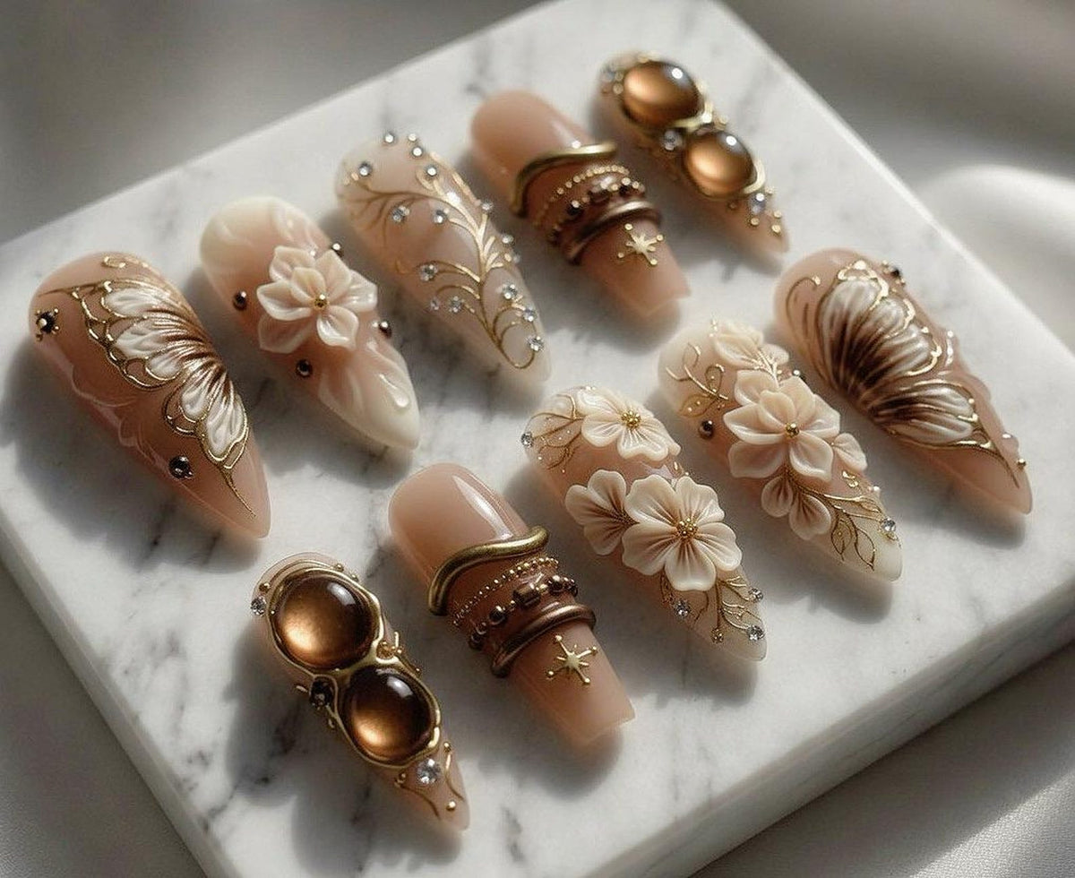 A collection of ten almond-shaped artificial nails. Each nail features intricate designs, including floral accents, gold filigree, and embedded gemstones. The nails are primarily nude with gold, brown, and white details, creating an elegant aesthetic