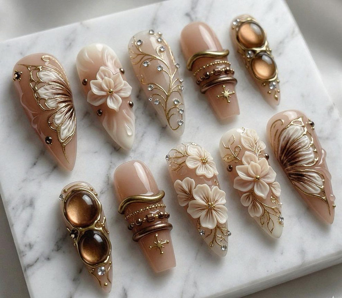 A collection of ten artificial nails with various designs. The nails feature a nude base color with intricate embellishments, including gold filigree, rhinestones, and 3D floral accents. Some nails have a butterfly wing design.
