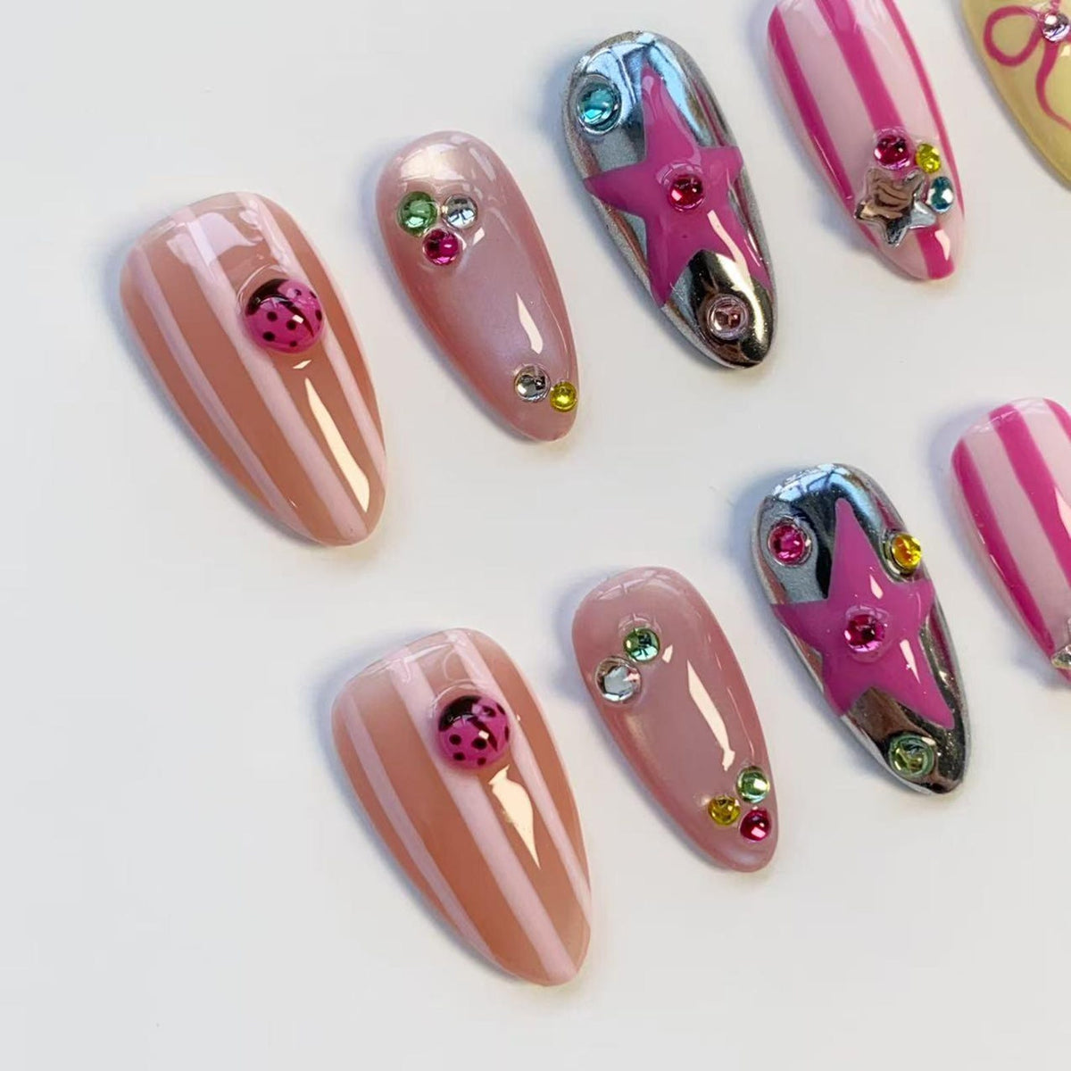A collection of artificial nails in various designs. Some nails are pink with white stripes, others are solid pink, and some feature silver chrome with pink star accents. Several nails are adorned with rhinestones and ladybug embellishments.