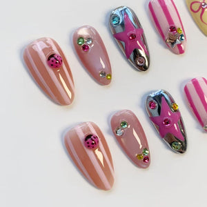 A collection of artificial nails in various designs. Some nails are pink with white stripes, others are solid pink, and some feature silver chrome with pink star accents. Several nails are adorned with rhinestones and ladybug embellishments.