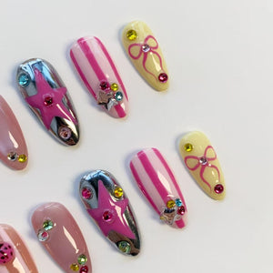 A collection of artificial nails in various designs. Some nails are pink with white stripes, others are chrome with pink stars, and some are yellow with pink bows. Each nail is adorned with colorful rhinestones.