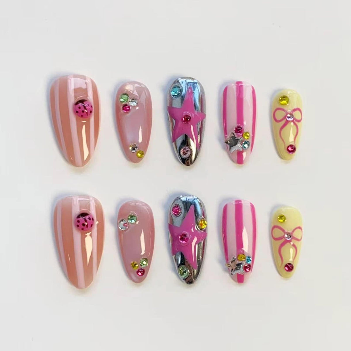 A set of ten artificial nails in various designs. Some nails feature pink and white stripes, ladybug accents, or colorful rhinestones. Others have a silver chrome finish with a pink star, or a yellow base with a pink bow.