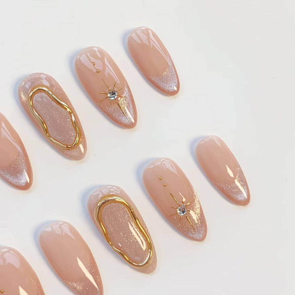 A set of almond-shaped press-on nails featuring a nude base with shimmering silver tips. Select nails are adorned with raised gold metallic swirls, while others feature a delicate gold starburst design with a small embedded clear crystal.