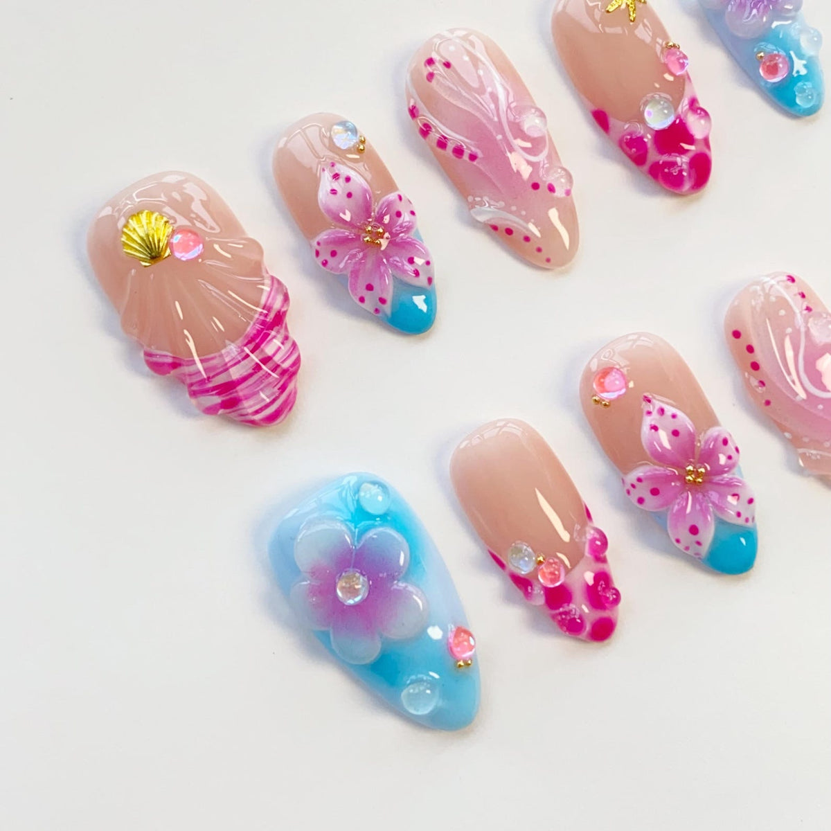 A collection of artificial nails with various designs. Some nails feature a gradient of nude to pink, adorned with pink flowers, gems, and blue tips. Others showcase seashell and wave patterns in pink and white, with gold accents. The nails are arran