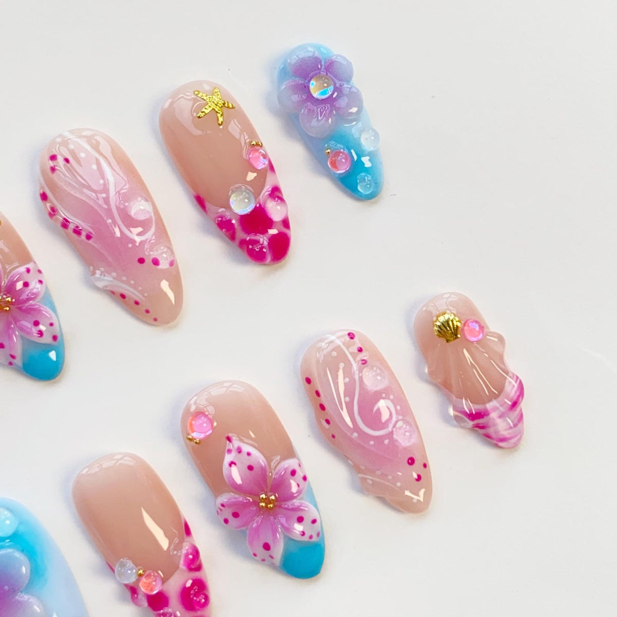 A collection of artificial nails with various designs. Some nails feature a nude base with pink and white floral accents, while others showcase blue and pink gradients with 3D flower and seashell embellishments. Gold star and gem accents are also pre