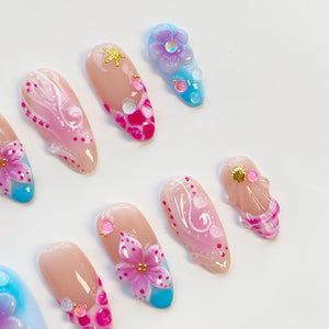 A collection of artificial nails with various designs. Some nails feature a nude base with pink and white floral accents, while others showcase blue and pink gradients with 3D flower and seashell embellishments. Gold star and gem accents are also pre