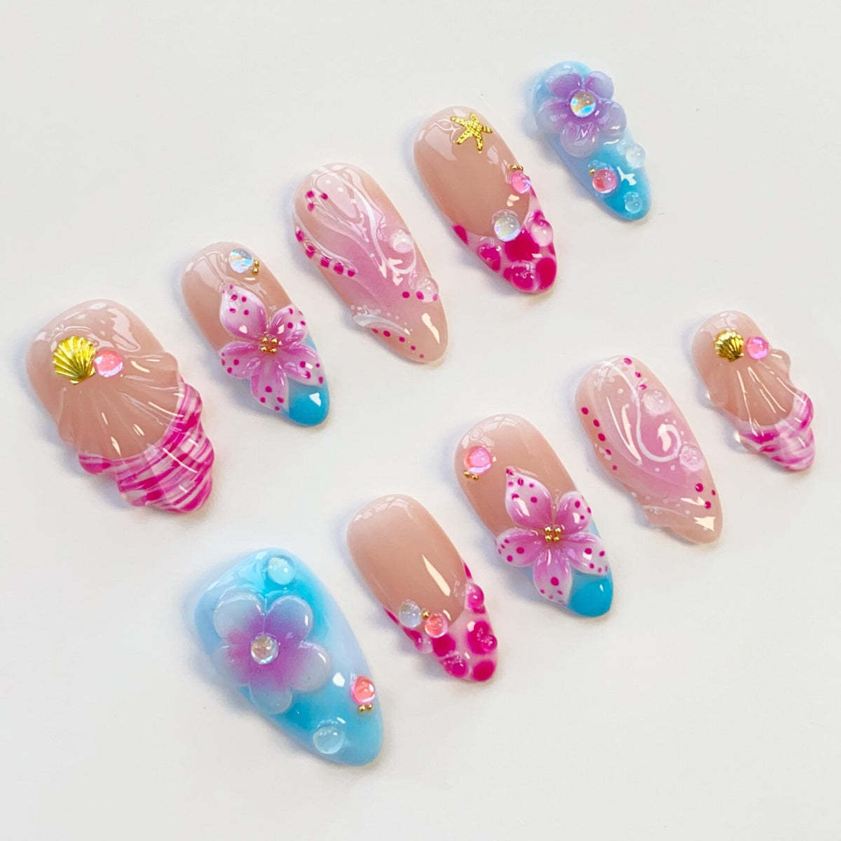 A collection of ten artificial nails with various designs. The nails feature a nude base with pink, blue, and white accents. Some nails have 3D flower and seashell embellishments, while others have painted patterns and rhinestones.