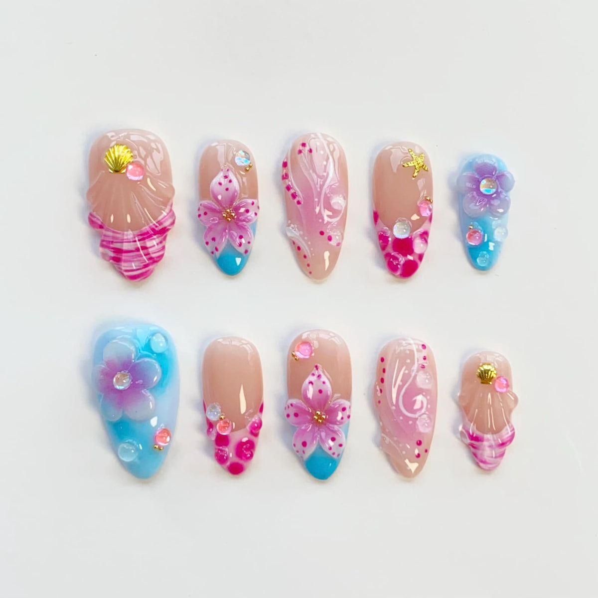 A set of ten artificial nails in various designs. The nails feature a nude base with pink, blue, and gold accents. Designs include flowers, seashells, and abstract patterns. The nails are oval-shaped.