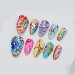 A set of ten press-on nails featuring vibrant, multi-colored marbled patterns, metallic silver star and crescent moon accents, and small embedded rhinestones in various shapes and colors, arranged in two rows on a white surface.