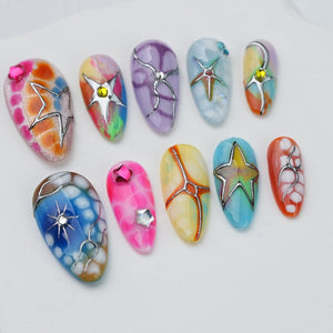 A set of ten press-on nails featuring various colorful designs, including marbled patterns, bright pink, blue, and orange hues, accented with silver metallic star-shaped charms, thin silver lines, and small embedded rhinestones.