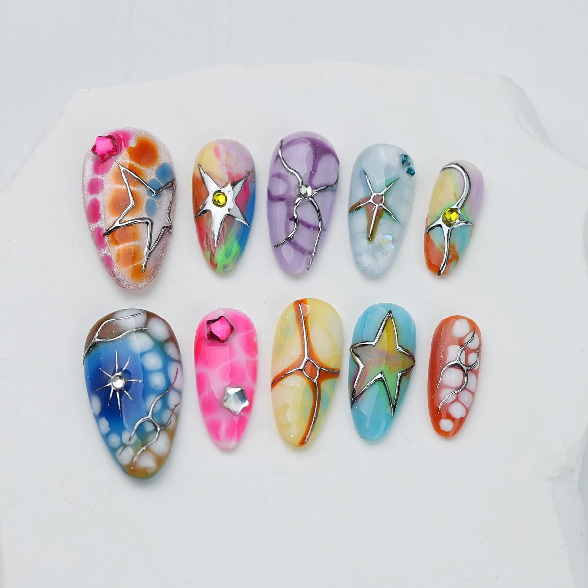 A set of ten press-on nails featuring various colorful designs, including abstract patterns, gradients, and marble effects. Several nails are decorated with metallic silver star-shaped accents, small rhinestones, and intricate silver line work.
