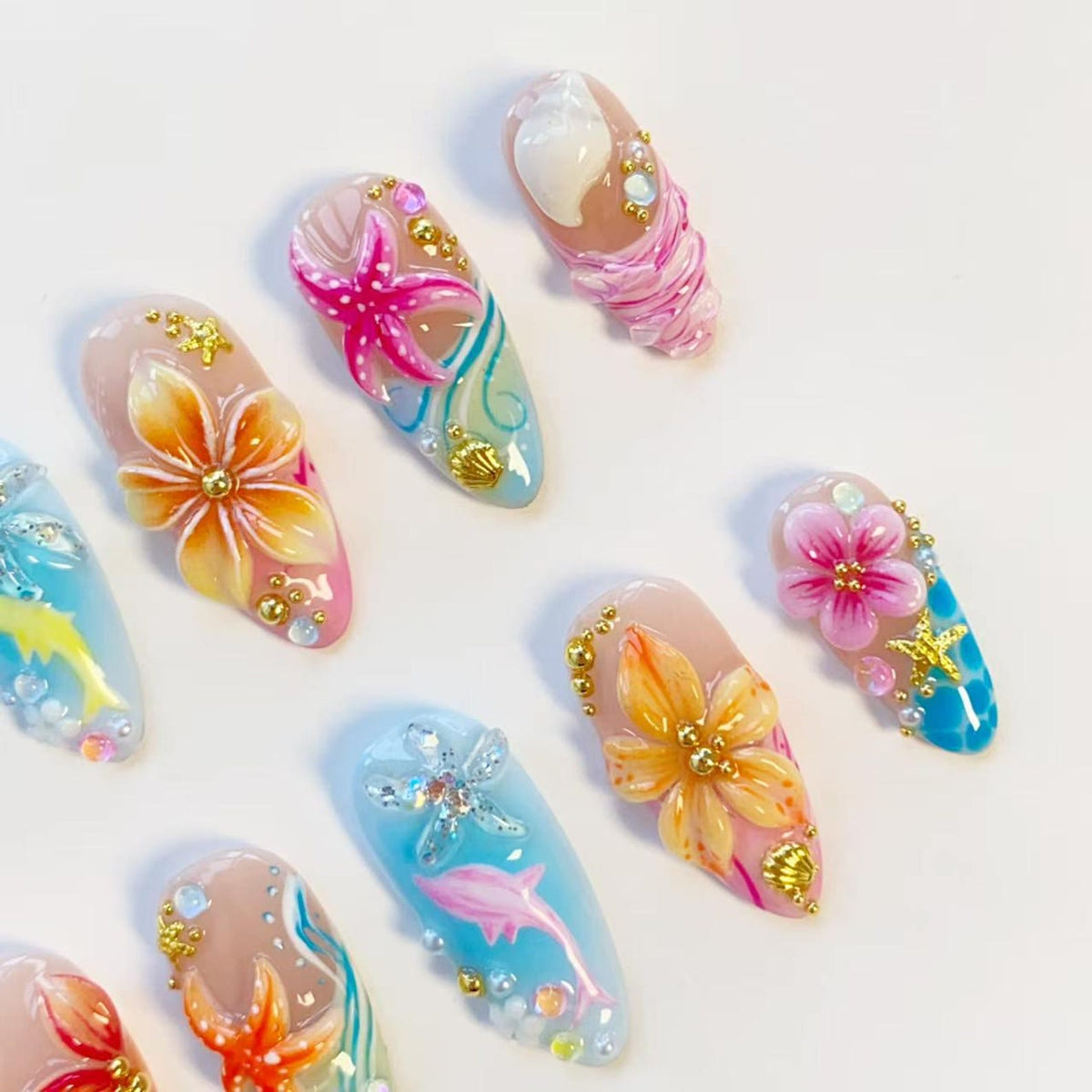 A collection of press-on nails featuring 3D designs with tropical themes. The nails display hand-painted flowers, starfish, and dolphins in shades of pink, orange, and blue, accented with gold beads, small seashells, and clear rhinestones.