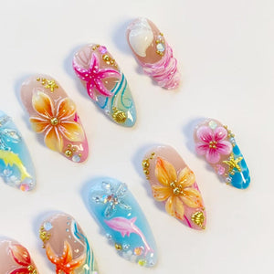 A collection of press-on nails featuring 3D designs with tropical themes. The nails display hand-painted flowers, starfish, and dolphins in shades of pink, orange, and blue, accented with gold beads, small seashells, and clear rhinestones.