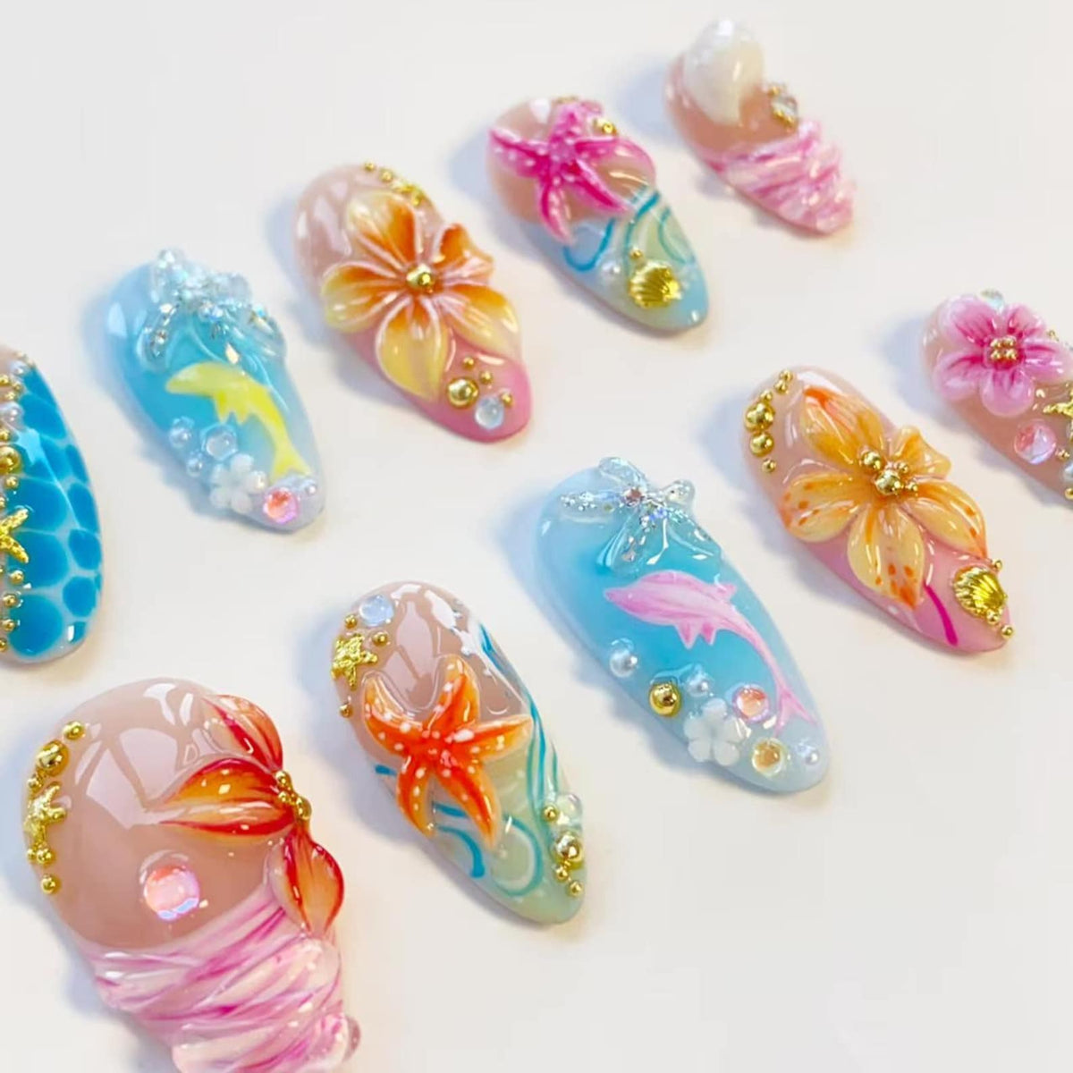 A collection of almond-shaped press-on nails featuring 3D designs including orange and pink flowers, starfish, dolphins, and seashells. The nails are decorated with small gold beads, clear rhinestones, and swirling blue and pink paint.