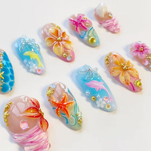 A collection of almond-shaped press-on nails featuring 3D designs including orange and pink flowers, starfish, dolphins, and seashells. The nails are decorated with small gold beads, clear rhinestones, and swirling blue and pink paint.