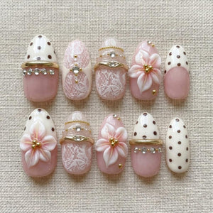 A set of ten almond-shaped press-on nails featuring a soft pink and cream color palette. Designs include white lace patterns, brown polka dots, three-dimensional pink flower appliqués, gold-toned metal bands, and clear rhinestones.