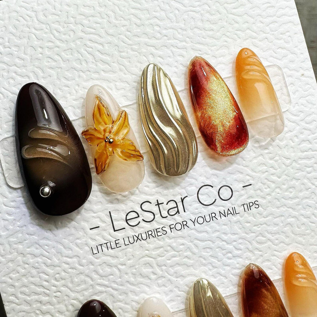 A set of five almond-shaped press-on nails featuring varied designs including a dark brown nail with a silver stud, a cream nail with a 3D flower, a metallic gold wavy nail, a red and gold cat-eye nai