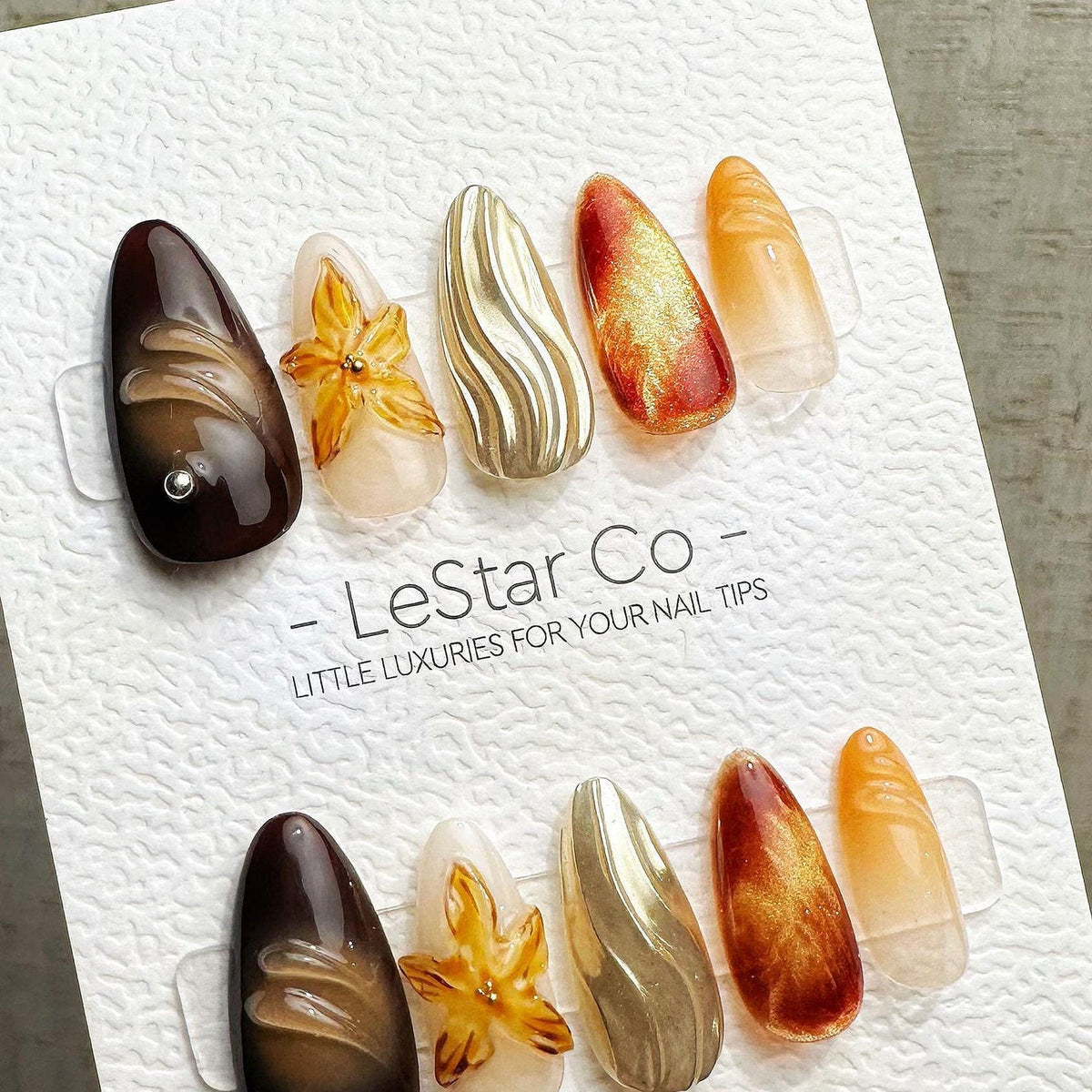 A set of ten almond-shaped press-on nails on a white display card labeled LeStar Co - LITTLE LUXURIES FOR YOUR NAIL TIPS. The designs include dark brown, translucent floral, metallic gold wavy, and am
