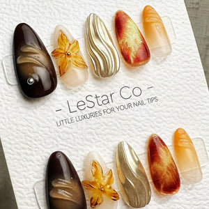 A set of ten almond-shaped press-on nail tips arranged on a white textured card labeled LeStar Co. The designs include dark brown with silver accents, translucent yellow flowers, metallic gold waves, 