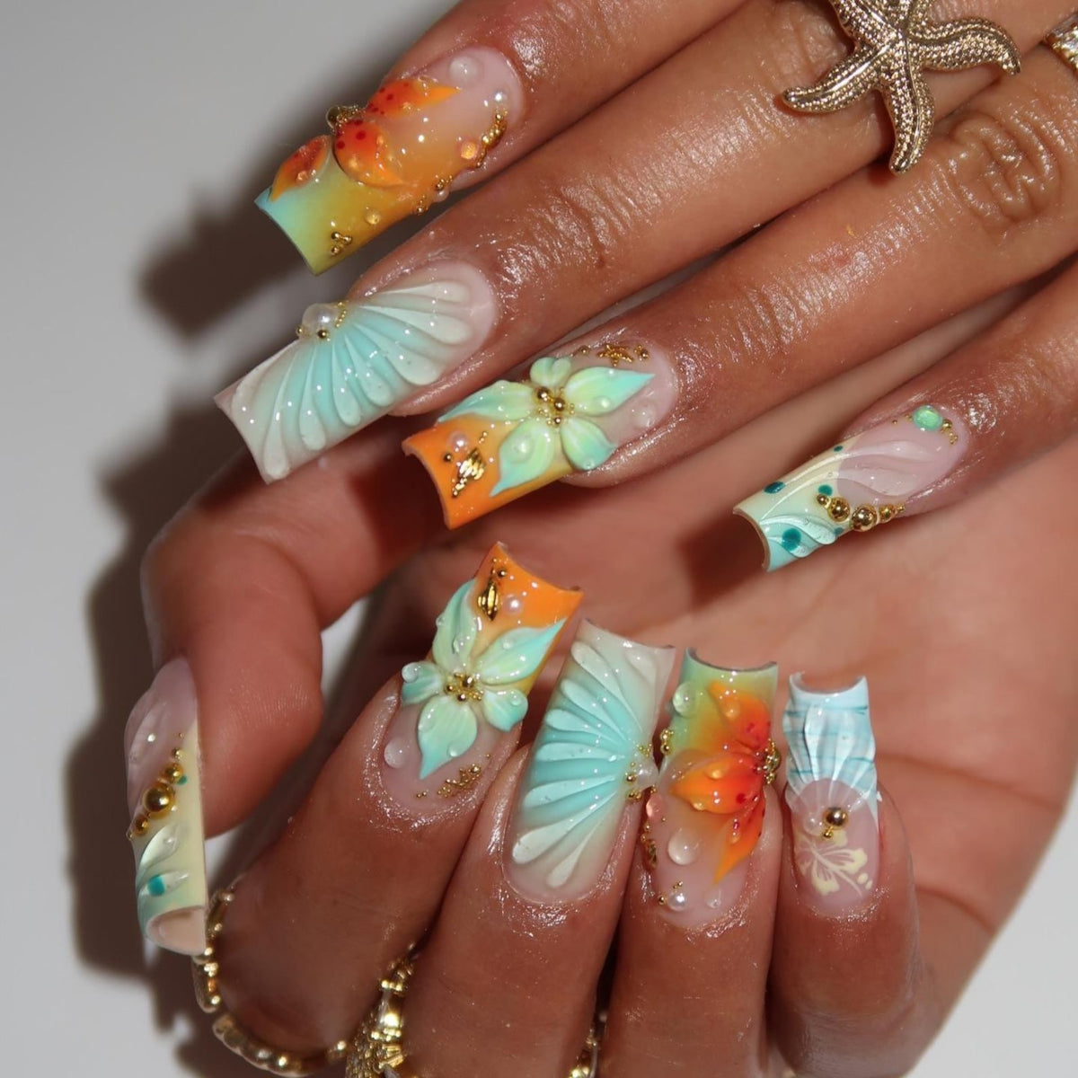 Long square-shaped artificial nails featuring a gradient of orange and light blue. The nails are decorated with three-dimensional sculpted flowers, raised shell patterns, small gold metallic beads, and clear water droplet accents.