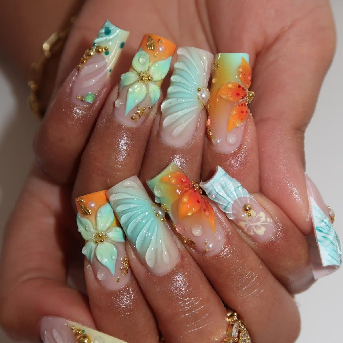 Square-shaped artificial nails featuring a gradient of orange and mint green. The design includes three-dimensional sculpted flowers, ribbed shell-like textures, small gold metallic beads, and clear droplet accents over a nude base.